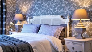 Ein Schlafzimmer mit weißem Kopfteil, zwei Nachttischen mit Lampen, einem Bett mit vielen Kissen und einem floralen Wandmuster.