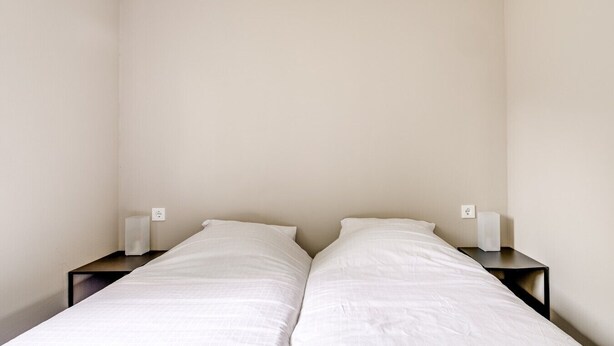Una habitación sencilla y sin adornos con dos camas individuales, cada una con colchón y almohada blancos, flanqueadas por mesitas de noche oscuras.