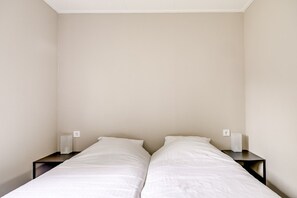 Una habitación sencilla y sin adornos con dos camas individuales, cada una con colchón y almohada blancos, flanqueadas por mesitas de noche oscuras.