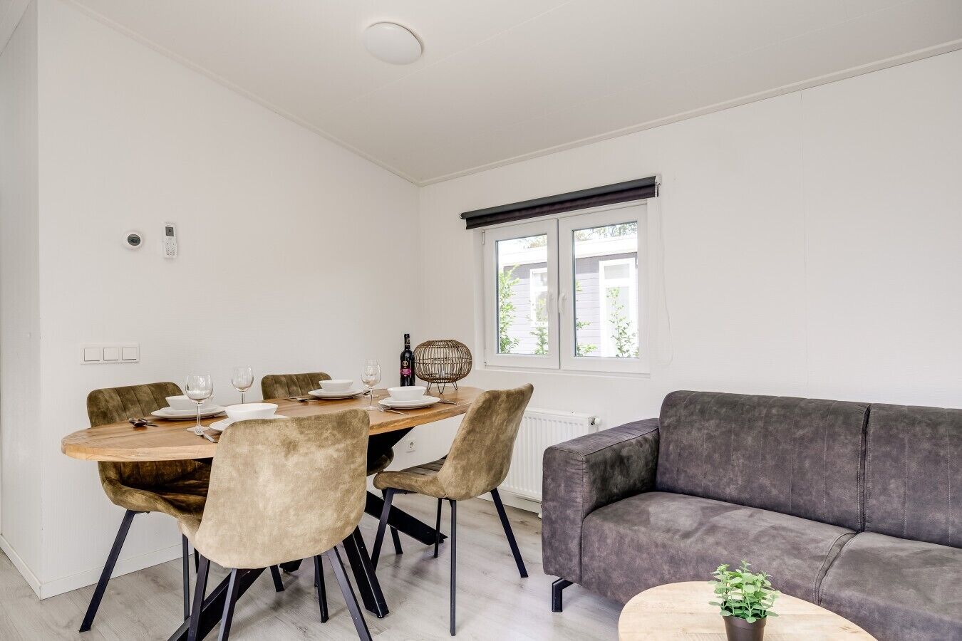Un coin repas avec une table en bois, des chaises rembourrées, un canapé gris et une fenêtre avec des stores.