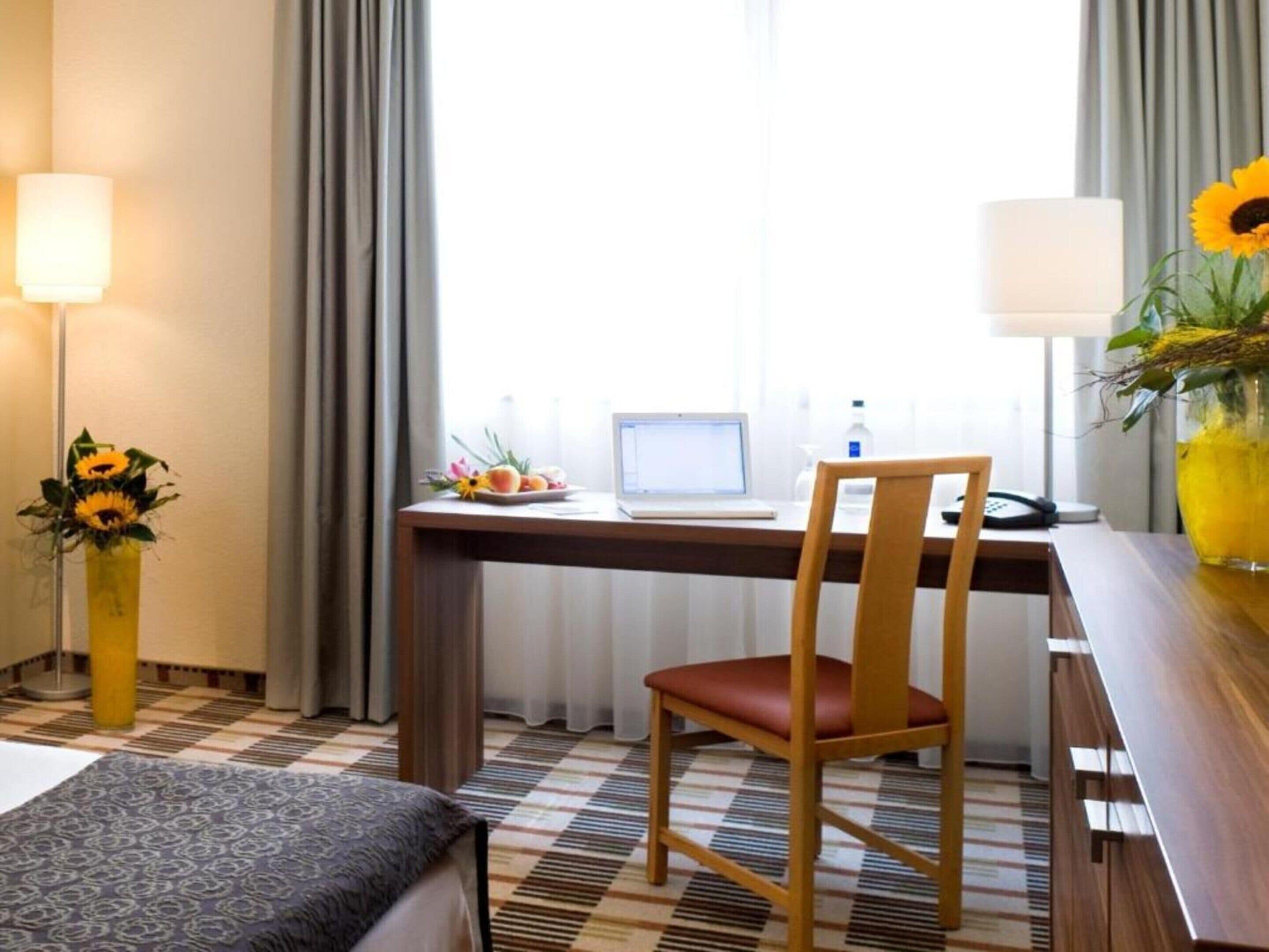 Habitación de hotel con cama, escritorio con ordenador portátil, silla, jarra con girasoles y lámpara.