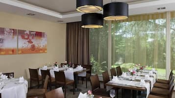 Un comedor con mesas dispuestas para el servicio, amplias ventanas con vistas a la vegetación e iluminación colgante moderna.