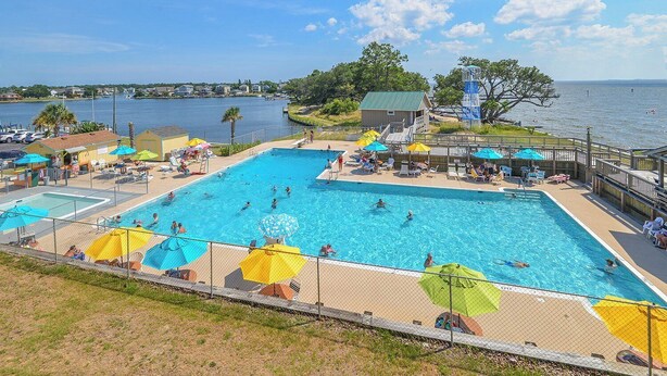 Uma grande piscina ao ar livre com várias pistas de natação, cercada por guarda-sóis e espreguiçadeiras, com vista para a água e edifícios ao longe.