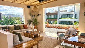 Ein modernes Wohnzimmer mit Sofa, Couchtisch und Esstisch, großen Fenstern und Blick auf ein Gebäude mit Balkonen.