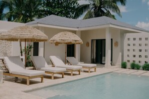 A poolside area with lounge chairs and thatched umbrellas, a stone wall, and a palm tree in the background. - Luxury 5BR Villa Pool, Jacuzzi, Rooftop Near Beach (Las Terrenas)
