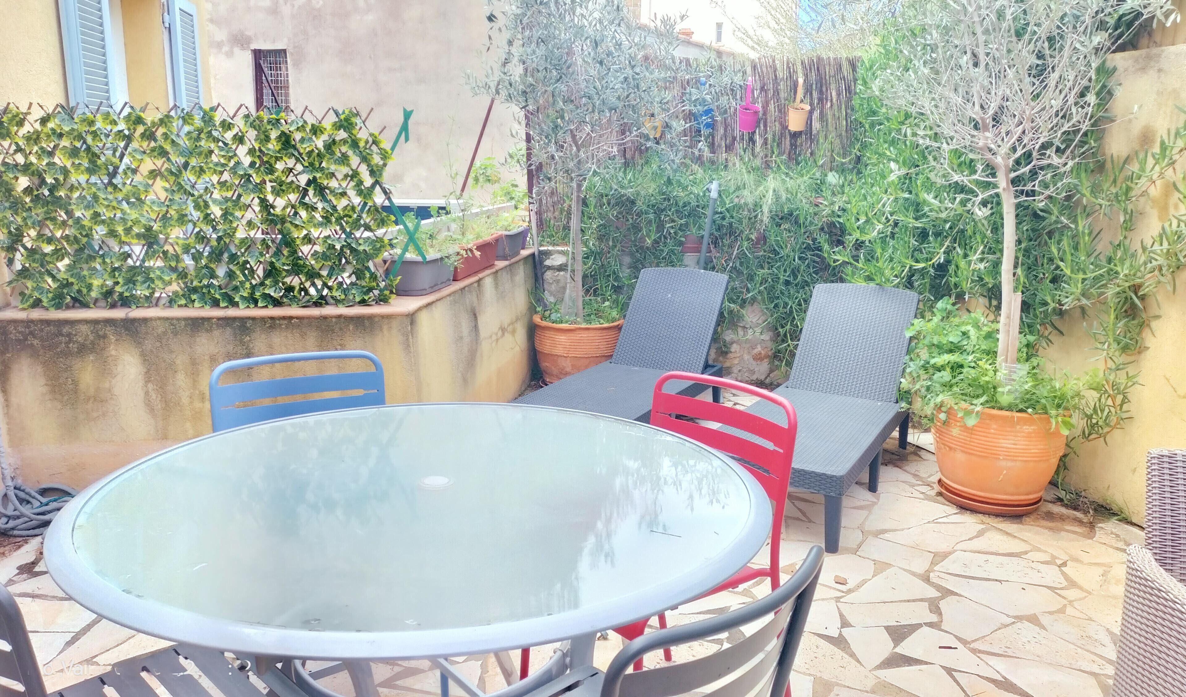 Une terrasse extérieure avec une table ronde en verre, deux chaises et des plantes en pot.