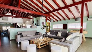 Uma sala de estar espaçosa com pé direito alto, vigas de madeira, um conjunto de sofás, uma mesa de centro e uma área de refeições ao fundo.