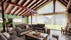Uma sala de estar ampla com um sofá seccional grande, uma mesa de centro de madeira e vista para o exterior por grandes janelas.