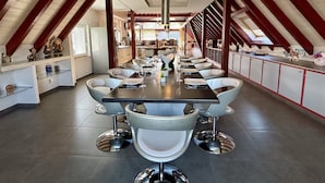 Uma sala de jantar com uma mesa comprida, cadeiras brancas e uma cozinha ao fundo.