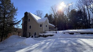 A two-story house with a stone chimney, a snow-covered driveway, and a forested backdrop. - Lovely 5-bedroom house in Londonderry perfect for your getaway (Londonderry)