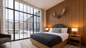 A modern bedroom with a large window, a bed with a dark headboard, a nightstand, and a wall-mounted deer head decoration. - LARCH HOUSE (Whitefish)