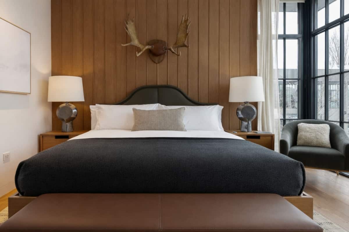 Ett hotellrum med en stor säng, två sänglampor, ett uppstoppat rådjurshuvud på väggen, ett fönster med utsikt och en bänk vid fotändan av sängen.