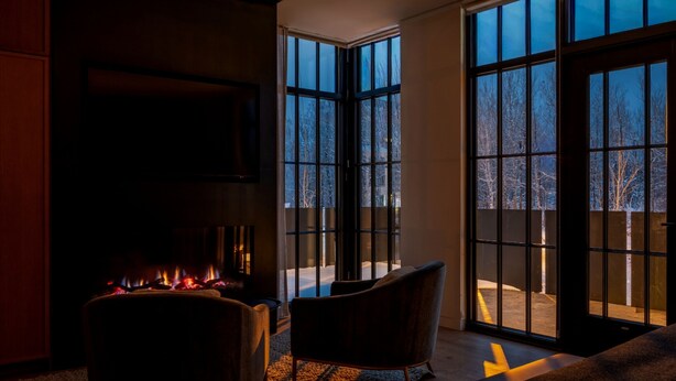 A room with a fireplace, two chairs, and large windows with a view of trees. - LARCH HOUSE (Whitefish)