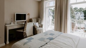 Une chambre d’hôtel avec un lit, un bureau équipé d’un ordinateur, une chaise et une vue sur un balcon donnant sur des immeubles.