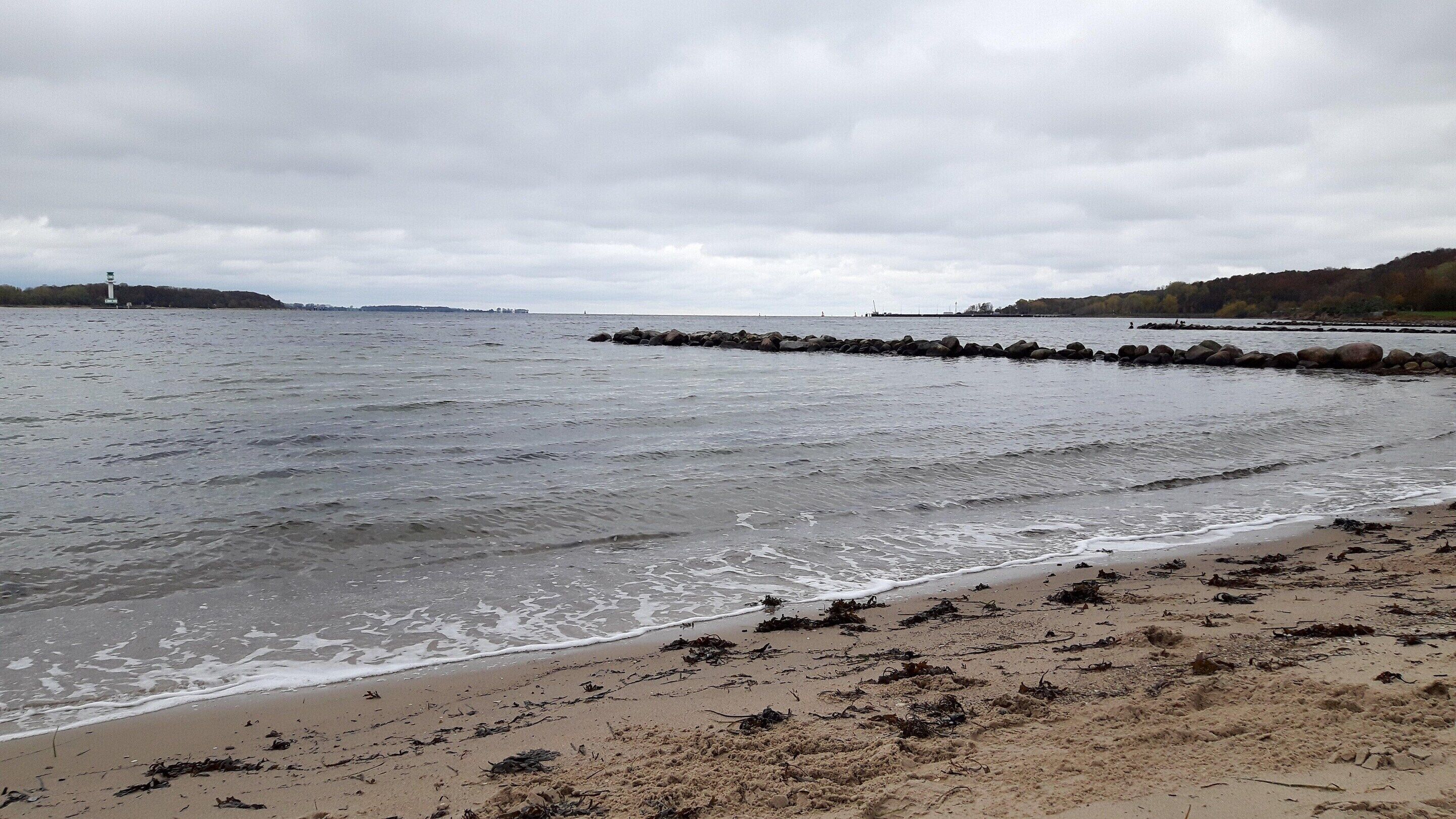 A beach with waves, a rocky breakwater, and a lighthouse in the distance.
