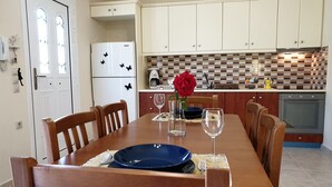Un coin repas avec une table en bois, des chaises, une assiette bleue, des verres Ă vin et un vase avec une fleur rouge.