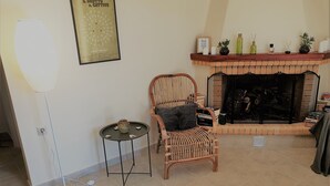 Un salon avec une cheminée, un fauteuil en osier, une petite table sur laquelle est posé un vase, et une affiche encadrée au mur.