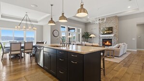 A spacious kitchen with a large island, pendant lighting, a stone fireplace, and a view of the outdoors. - Luxe Manor - Heated Lakefront Pool, Lily Pad & More! (Sunrise Beach)