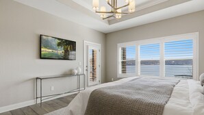 A bedroom with a bed, a painting, a door, and a window with blinds. - Luxe Manor - Heated Lakefront Pool, Lily Pad & More! (Sunrise Beach)