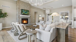 A modern living room with a stone fireplace, white sofas, a dining area, and a kitchen visible in the background. - Luxe Manor - Heated Lakefront Pool, Lily Pad & More! (Sunrise Beach)