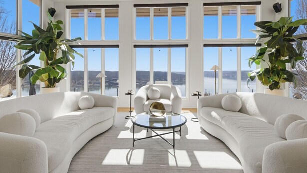 A spacious living room with large windows, two white sofas, a round glass coffee table, and potted plants.