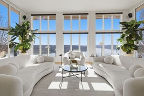 A spacious living room with large windows, two white sofas, a round glass coffee table, and potted plants. - Vantage Point - Heated Pool, Hot Tub, & Views! (Sunrise Beach)