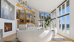 A modern living room with a large white sofa, a fireplace, and large windows offering a view of the outdoors.