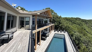 Una casa moderna con terraza de madera, piscina y balcón con vistas a una ladera boscosa.