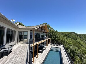 A modern house with a wooden deck, a pool, and a balcony overlooking a forested hillside.