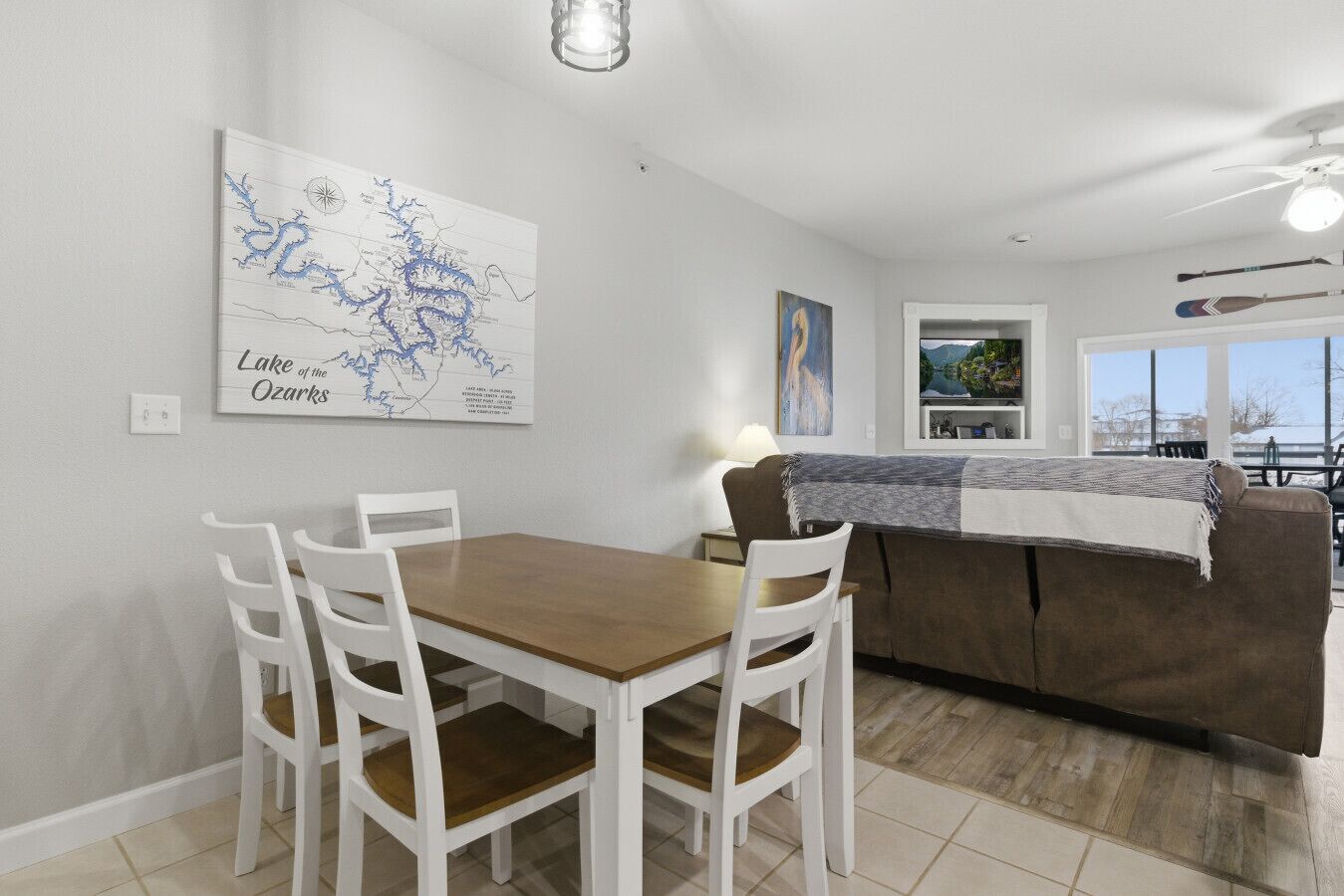 Uma área de refeições com uma mesa de madeira e cadeiras brancas, um sofá marrom com um cobertor, um mapa na parede e uma TV em uma estante.