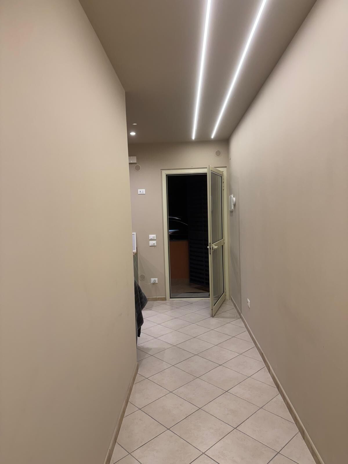 Un couloir étroit avec un sol carrelé, une porte donnant sur une pièce sombre et un éclairage encastré.
