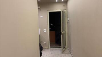 Un couloir étroit avec un sol carrelé, une porte donnant sur une pièce sombre et un éclairage encastré.