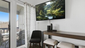 A room with a TV mounted on the wall, a wooden table, two chairs, and a view of a deck with outdoor furniture. - Lakeview for Two - Lakeside Pool & Top Floor! (Osage Beach)