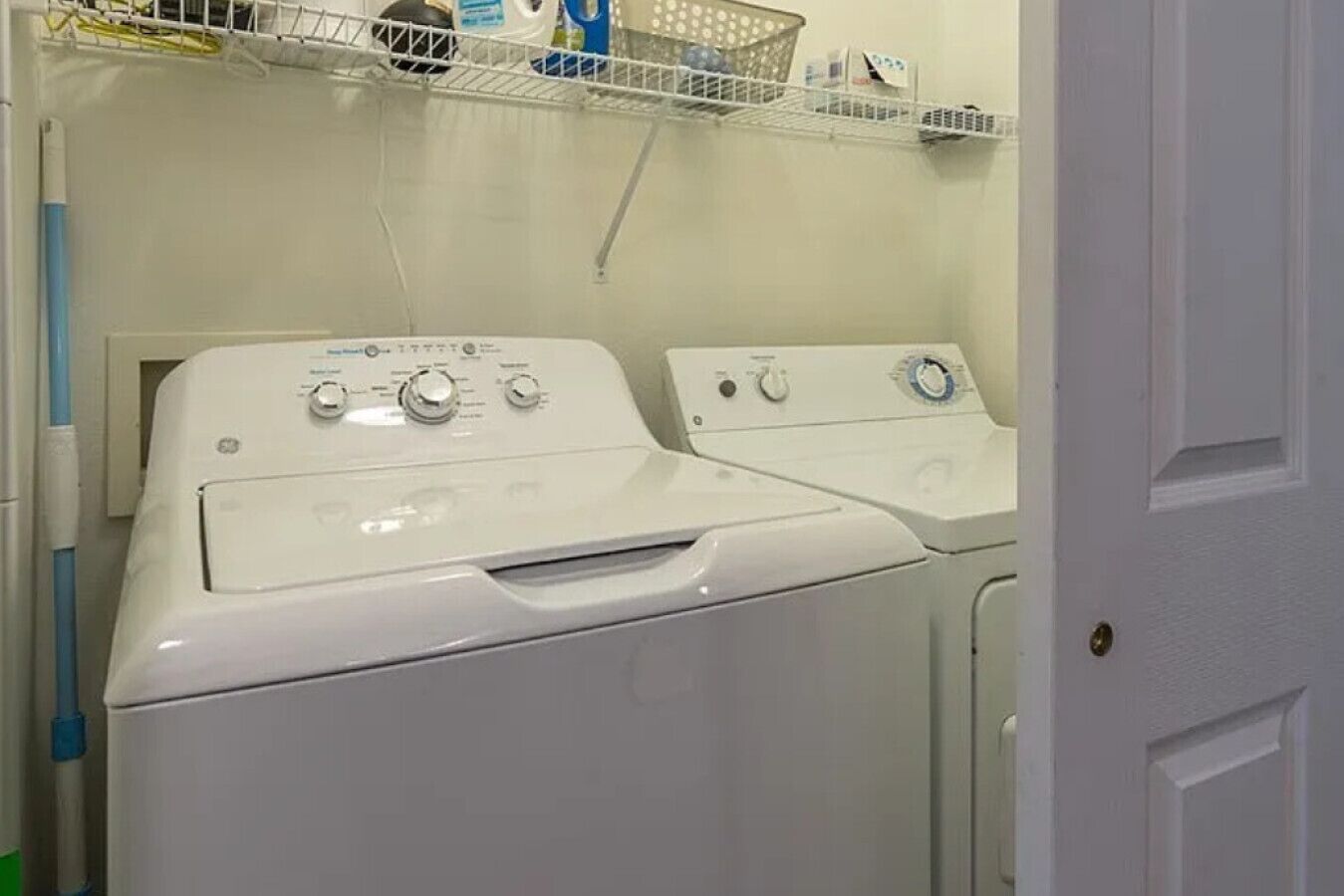 A laundry area with two front-loading washing machines, a shelf with laundry supplies, and a door to the right.