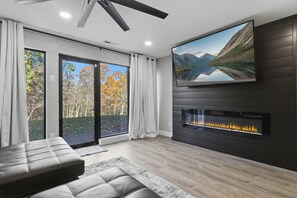 A modern living room with a fireplace, a large flat-screen TV, and a view of a forest through glass doors. - Graystone Getaway - Multiple pools! (Lake Ozark)