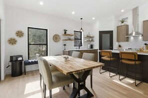 A modern kitchen with a dining area, wooden table, bar stools, and a large window.