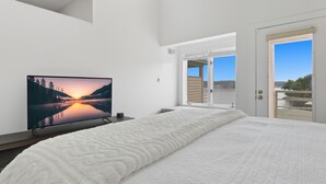 Een slaapkamer met een bed, een televisie waarop een zonsondergang te zien is, en een deur die uitkomt op een balkon met uitzicht op een waterpartij.
