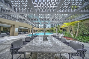 A poolside area with a long table and chairs, a pool, and a covered seating area with a geometric ceiling design. - Luxury 2BR/2BA w/ Pool & 50ft Balcony • 1mi to Beach • 8min to FLL (Dania Beach)