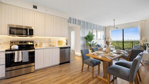 Una cocina moderna con electrodomésticos de acero inoxidable, un comedor con mesa y sillas de madera, y vistas a un paisaje urbano a través de grandes ventanales.
