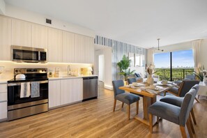 A modern kitchen with stainless steel appliances, a dining area with a wooden table and chairs, and a view of a cityscape through large windows. - Luxury 2BR/2BA w/ Pool & 50ft Balcony • 1mi to Beach • 8min to FLL (Dania Beach)