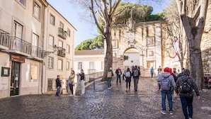 A cobblestone street with historic buildings, a stone archway, and people walking. - Nomad s Castel Lisbon - The Solo (Lisbon)