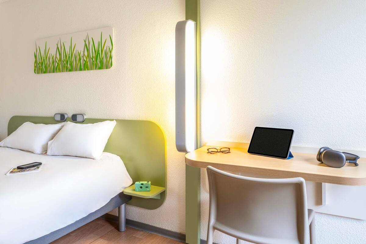 A hotel room with a bed, bedside table, chair, desk, and a wall-mounted lamp.