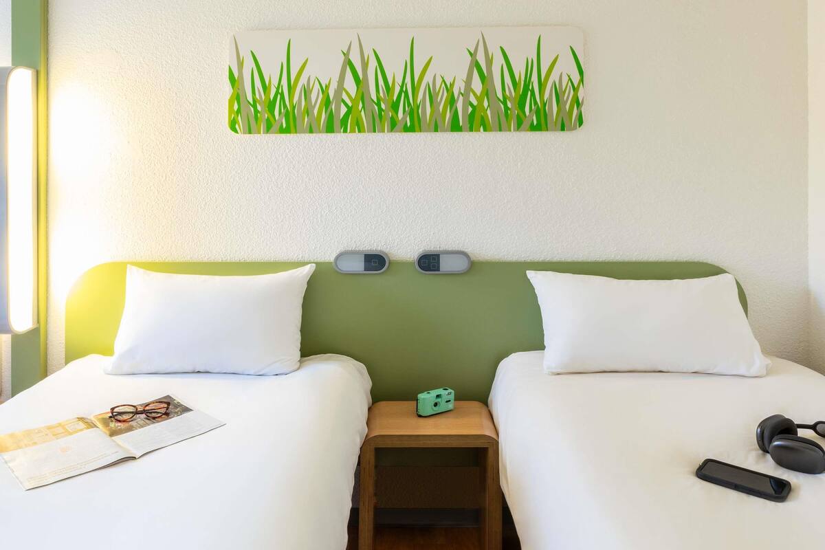 A hotel room with two beds, a small wooden nightstand, a wall-mounted digital clock, and a framed artwork of grass.
