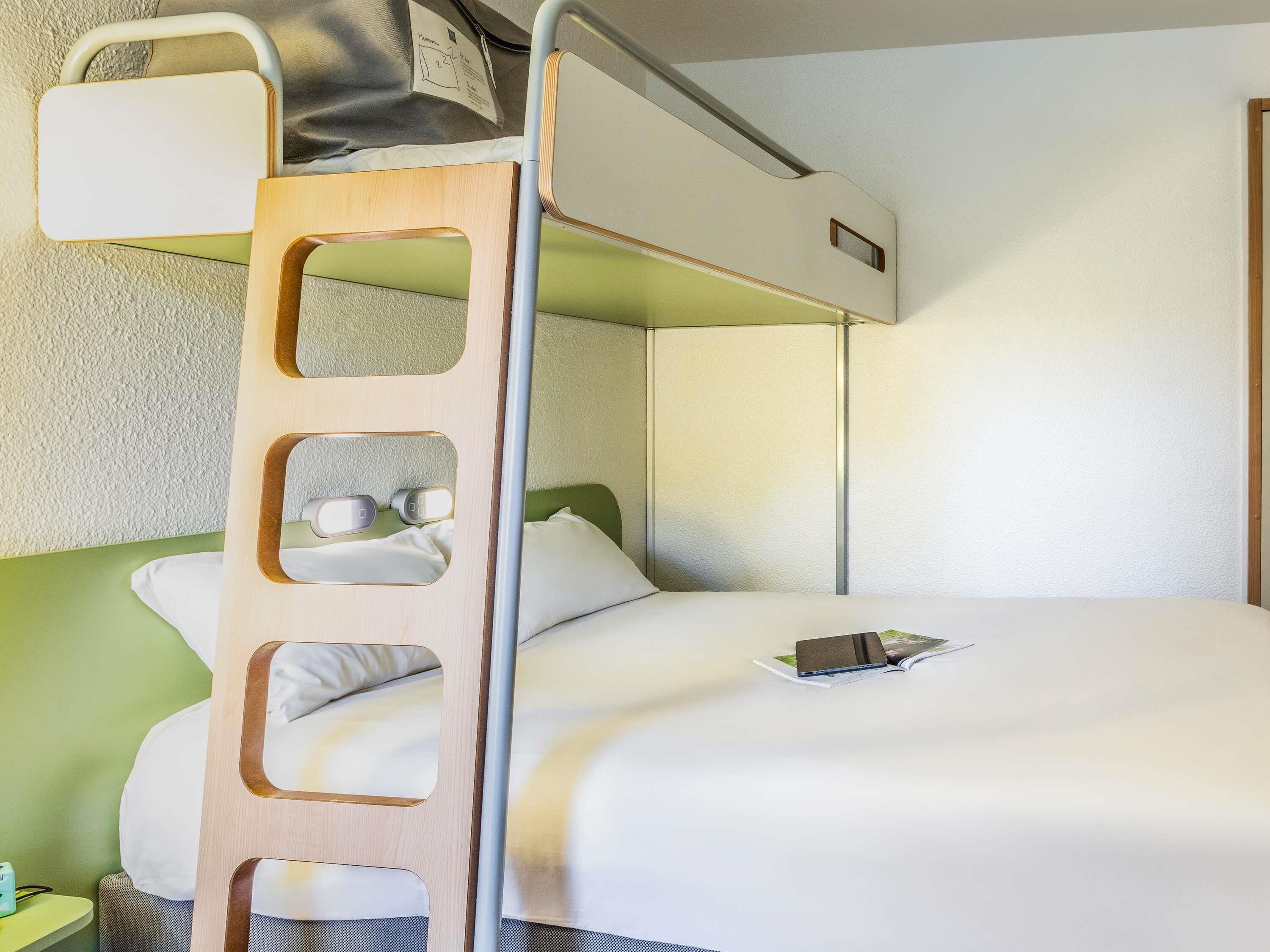 Una cama individual con escalera, una mesita de noche con lámpara, y una cama con almohada y un libro.