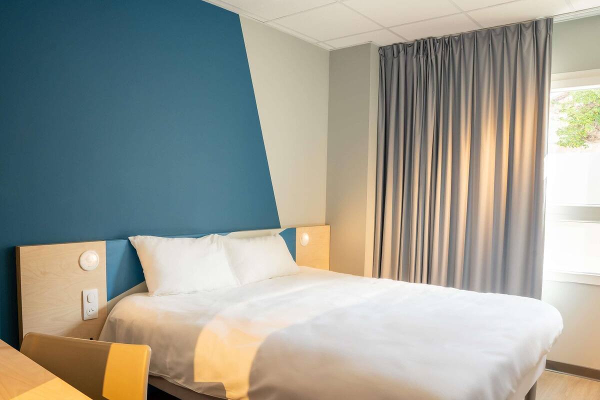 A hotel room with a bed, a bedside table, a wall with a blue panel, a window with curtains, and a thermostat on the wall.