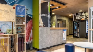 A hotel reception area with a reception desk, a waiting area with tables and chairs, and a wall with a map and information posters. - Ibis Budget Compiègne Centre-Ville (Compiegne)