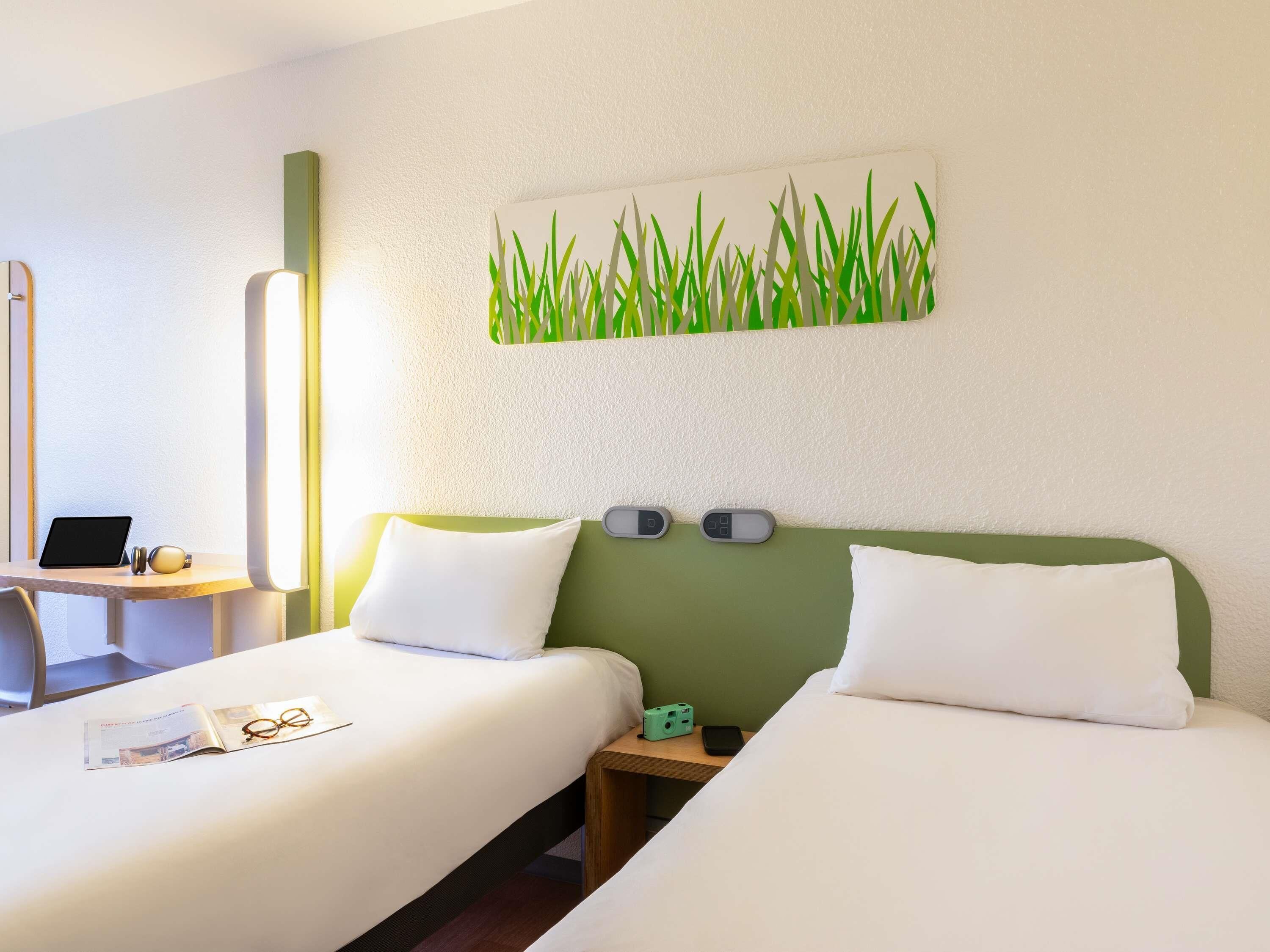 Une chambre d’hôtel avec deux lits, un bureau avec un ordinateur portable, une décoration murale verte et une table de chevet avec un téléphone et un appareil photo.