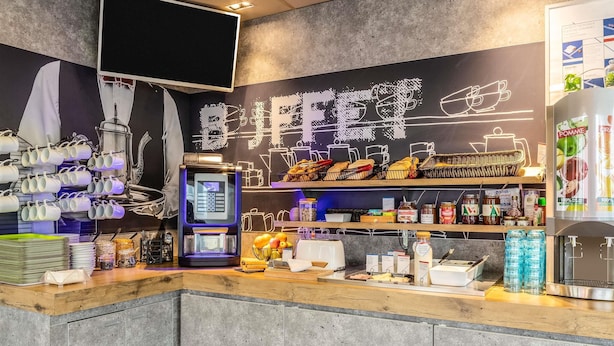 A café interior with a coffee machine, tea station, and shelves stocked with snacks and condiments. - Ibis Budget Compiègne Centre-Ville (Compiegne)