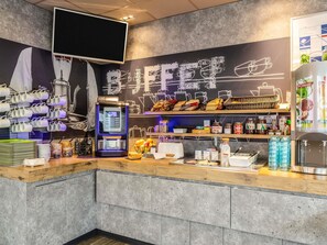 A café interior with a coffee machine, tea station, and shelves stocked with snacks and condiments. - Ibis Budget Compiègne Centre-Ville (Compiegne)