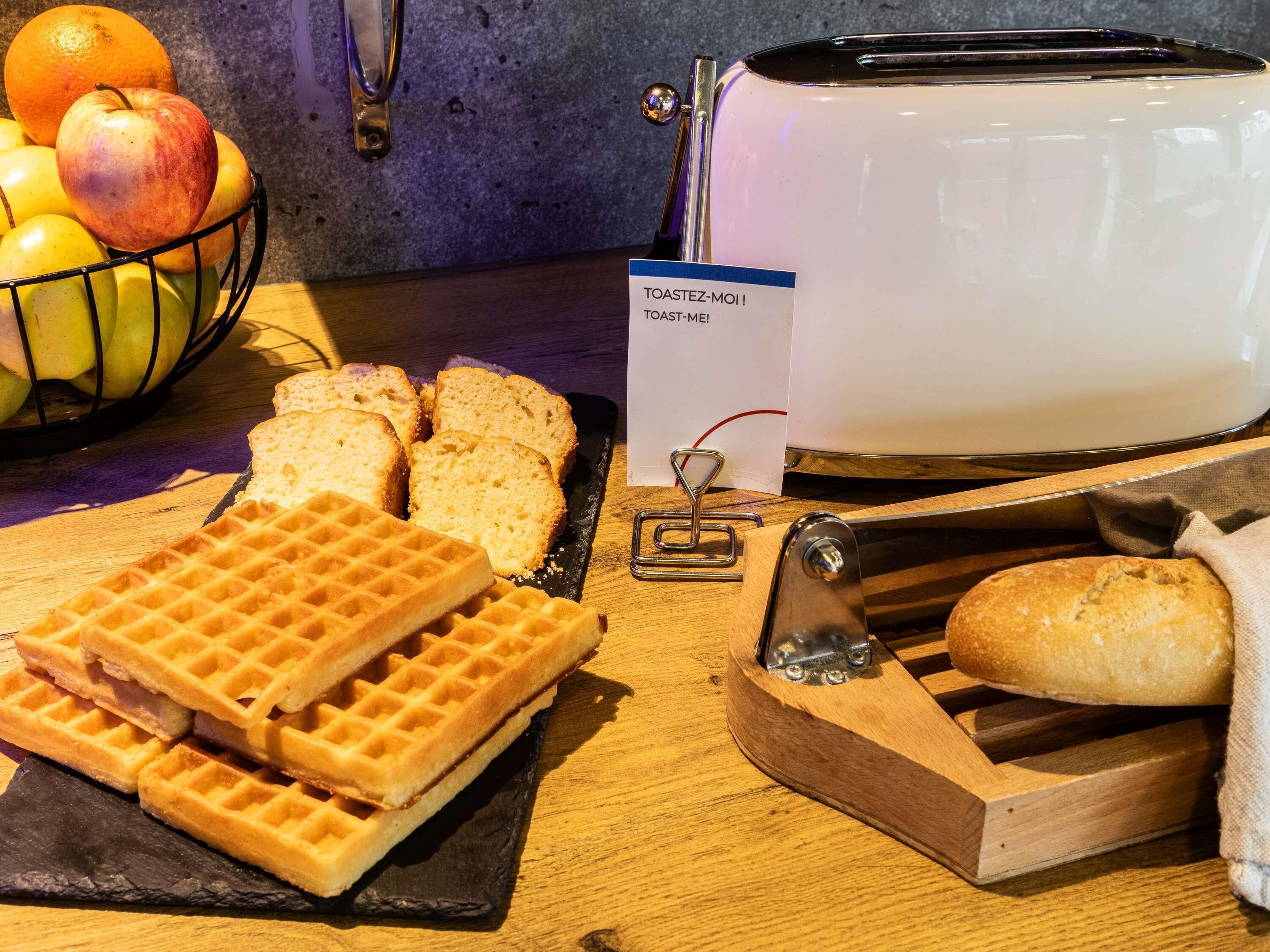 A kitchen counter with a toaster, a waffle maker, sliced bread, and a bowl of fruit.